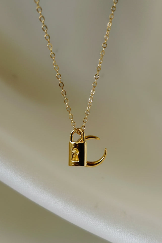 A delicate gold chain necklace with a small pendant featuring a black and gold geometric design, displayed on a white ceramic stand.