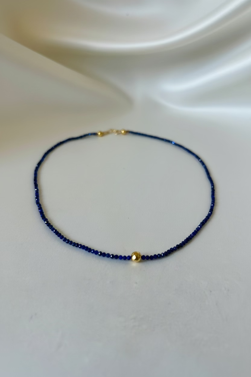A delicate beaded necklace with deep blue beads and small gold accents, displayed on a draped white satin fabric background.