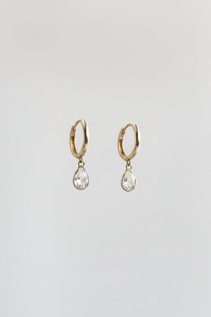 A pair of silver-toned huggie hoop earrings with dangling teardrop-shaped clear gemstones, displayed on a black metal stand against a warm beige and yellow draped background.
