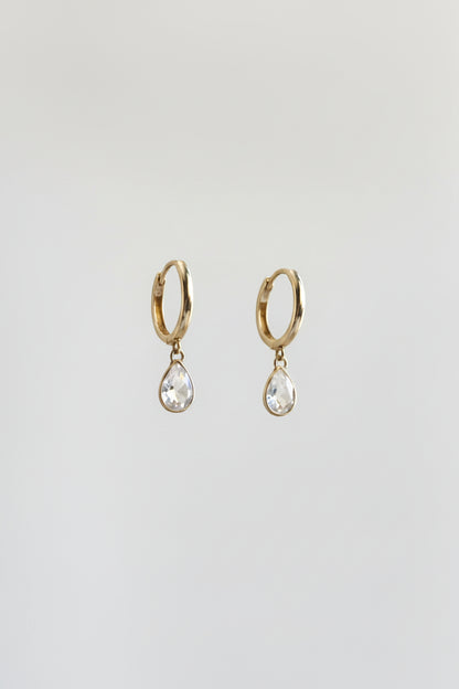 A pair of silver-toned huggie hoop earrings with dangling teardrop-shaped clear gemstones, displayed on a black metal stand against a warm beige and yellow draped background.
