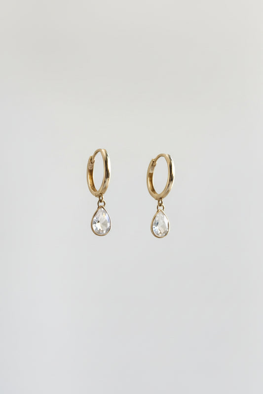 A pair of silver-toned huggie hoop earrings with dangling teardrop-shaped clear gemstones, displayed on a black metal stand against a warm beige and yellow draped background.