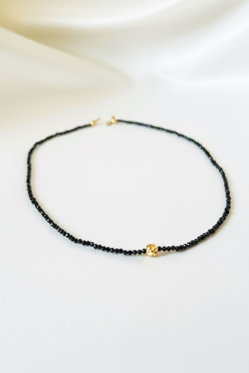 A delicate black beaded necklace with a small gold heart charm, displayed on a draped white satin fabric background.