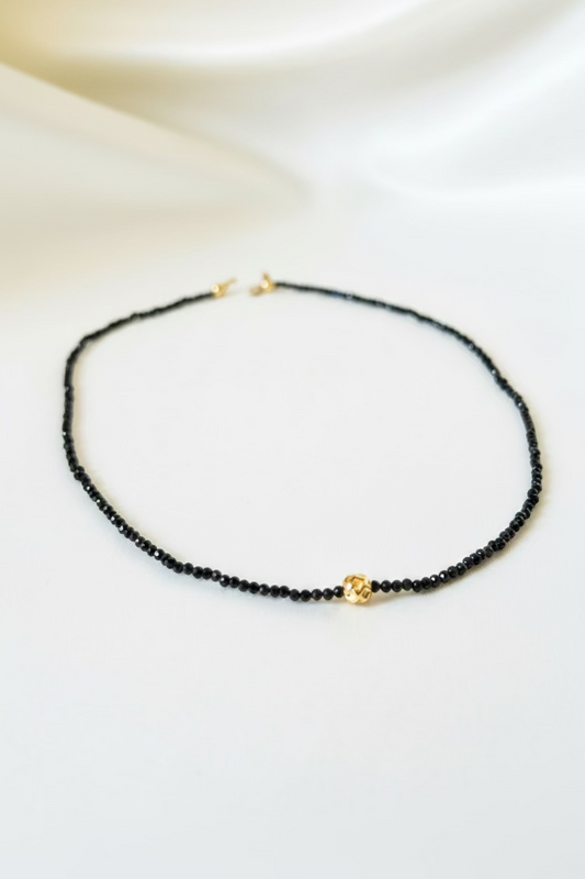 A delicate black beaded necklace with a small gold heart charm, displayed on a draped white satin fabric background.