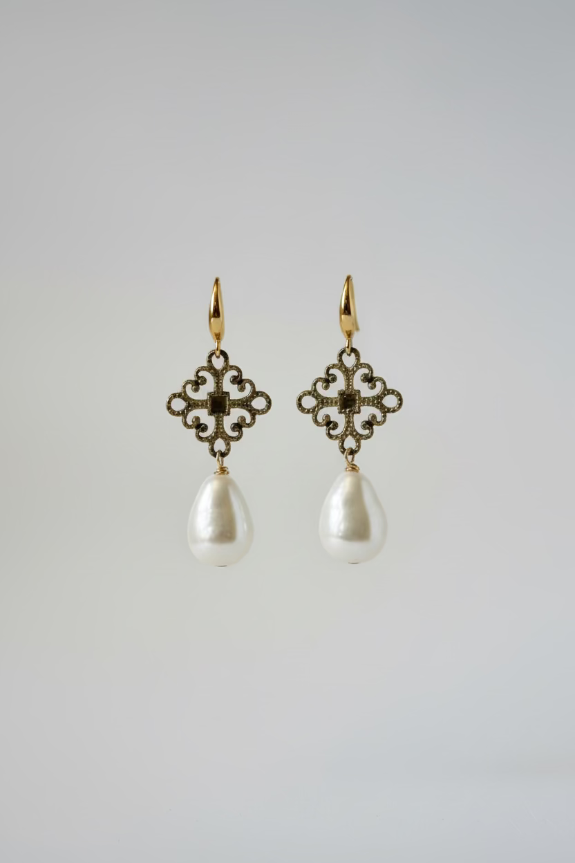 A pair of dangling earrings displayed on a metal stand against a draped satin background. Each earring has an ornate filigree-style gold-toned top with small clear stones and a large irregular white pearl drop below.