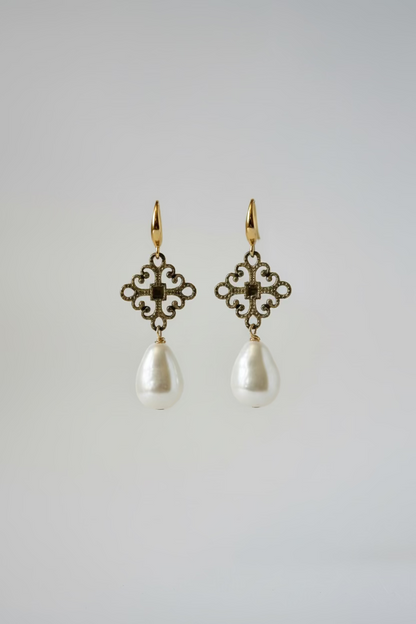 A pair of dangling earrings displayed on a metal stand against a draped satin background. Each earring has an ornate filigree-style gold-toned top with small clear stones and a large irregular white pearl drop below.