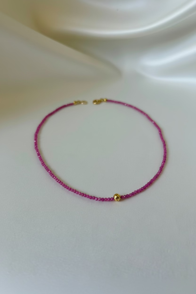 A delicate beaded necklace with deep red beads and small gold accents, including a central gold bead and clasp, displayed on a draped white satin fabric.