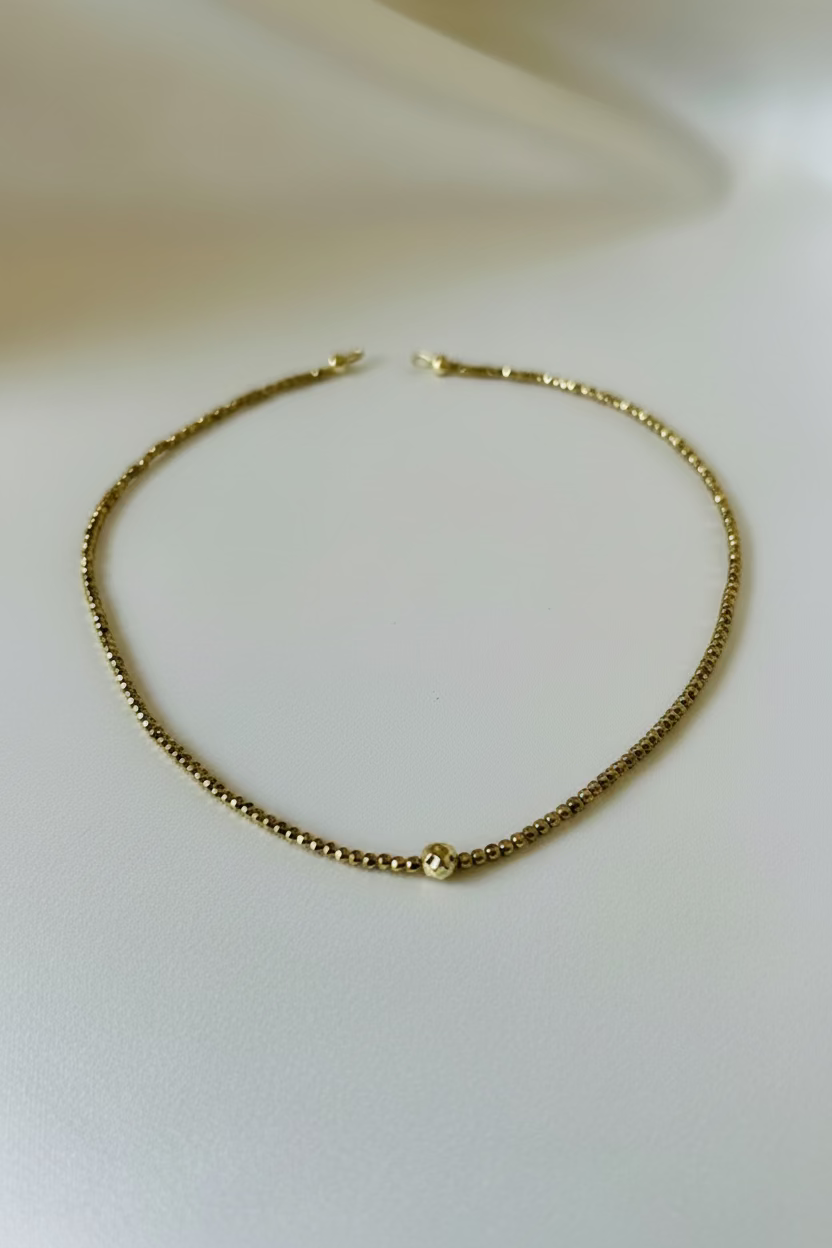 A delicate gold chain necklace with a small, round textured gold bead at the center, displayed on a soft, light-colored fabric surface.