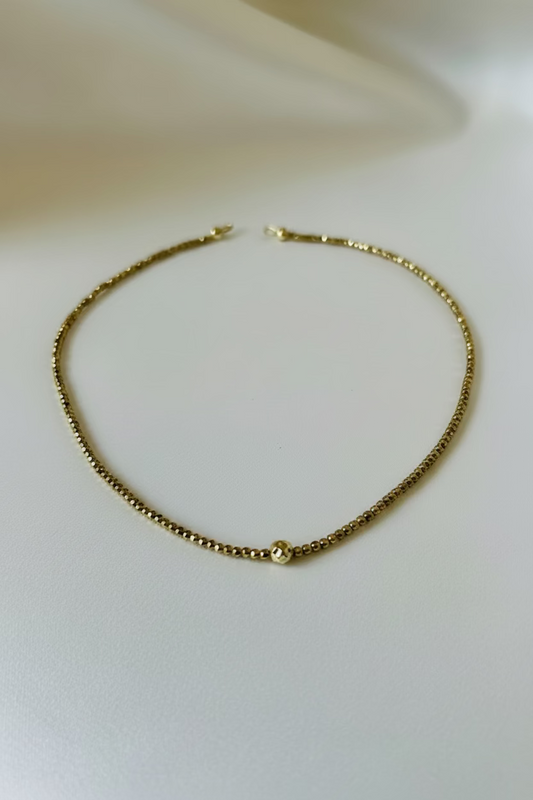 A delicate gold chain necklace with a small, round textured gold bead at the center, displayed on a soft, light-colored fabric surface.