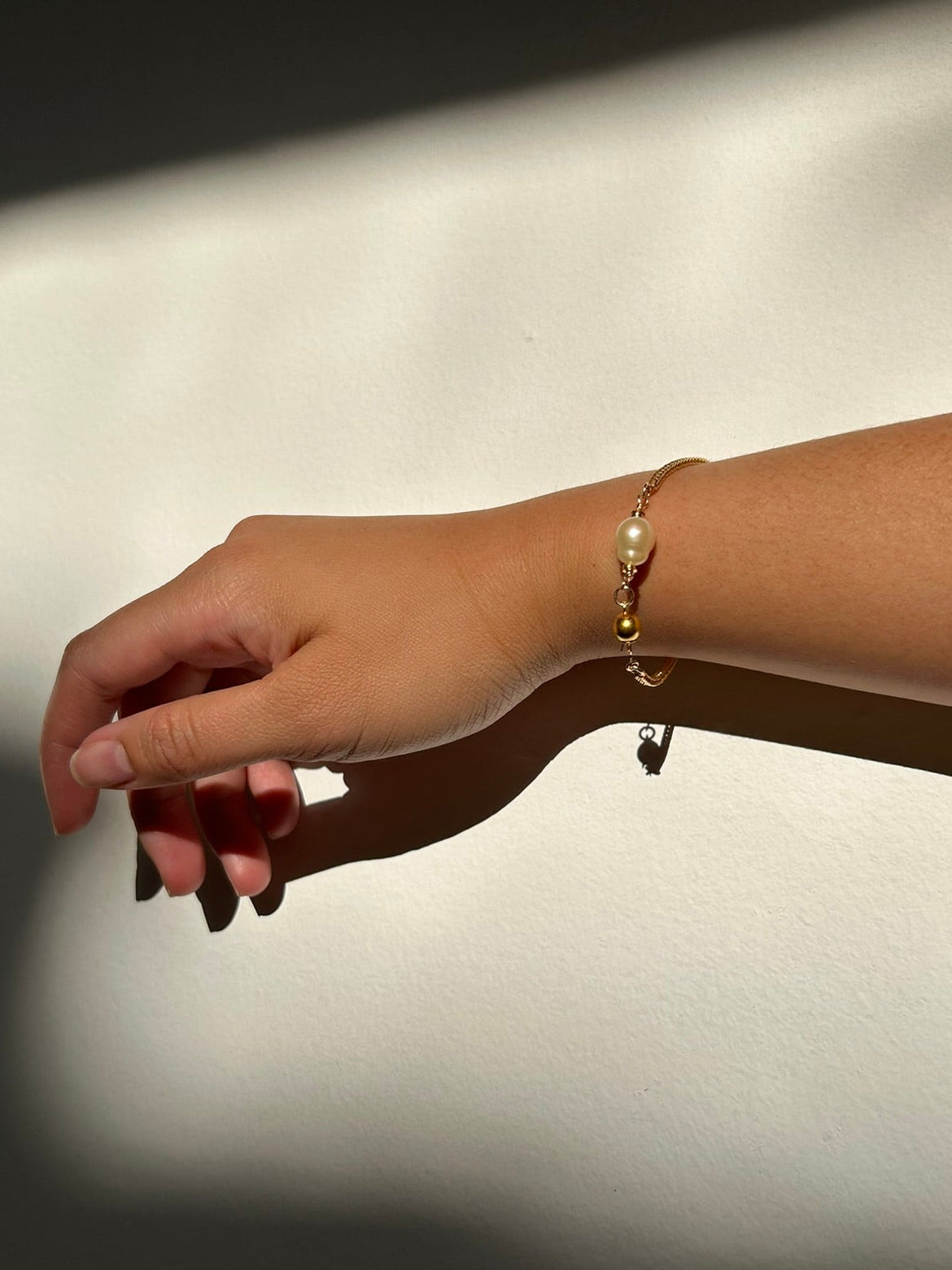 A delicate gold chain bracelet with a small white pearl and gold charm, worn on a person's wrist against a wall with dramatic diagonal shadows.