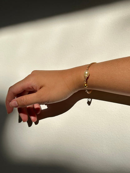A delicate gold chain bracelet with a small white pearl and gold charm, worn on a person's wrist against a wall with dramatic diagonal shadows.
