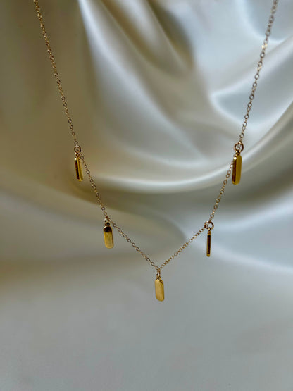 A delicate gold chain necklace with five small cylindrical bar pendants in varying lengths, displayed against a draped cream satin fabric background.