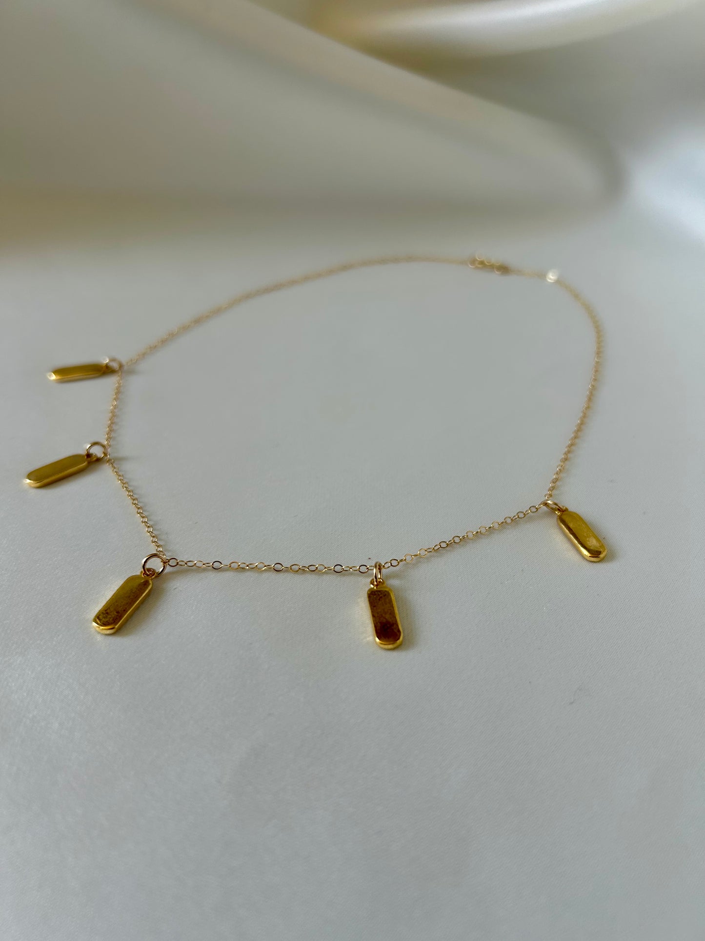 A delicate gold chain necklace with five small, elongated rectangular gold charms, displayed on a soft, light beige fabric background.