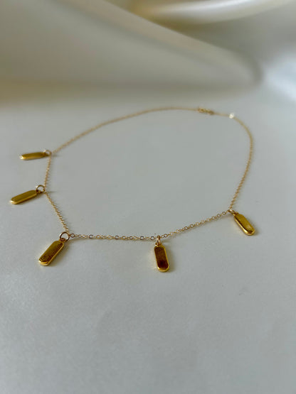 A delicate gold chain necklace with five small, elongated rectangular gold charms, displayed on a soft, light beige fabric background.