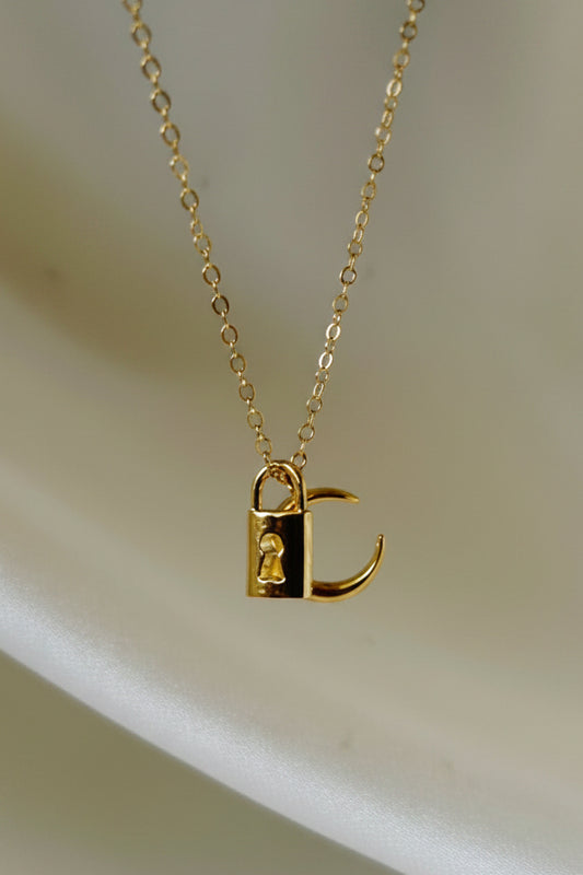 A delicate gold chain necklace with a small pendant featuring a black and gold geometric design, displayed on a white ceramic stand.