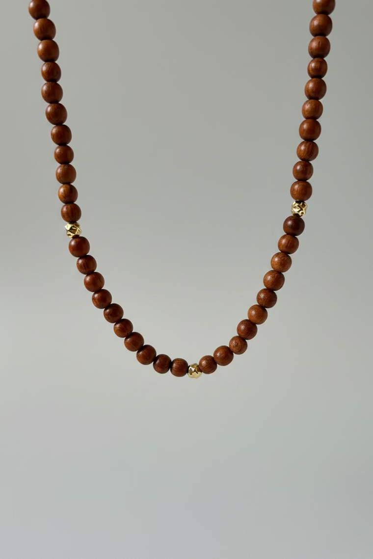 A beaded necklace made of dark reddish-brown round beads with small gold spacer accents, draped over a white satin display bust.