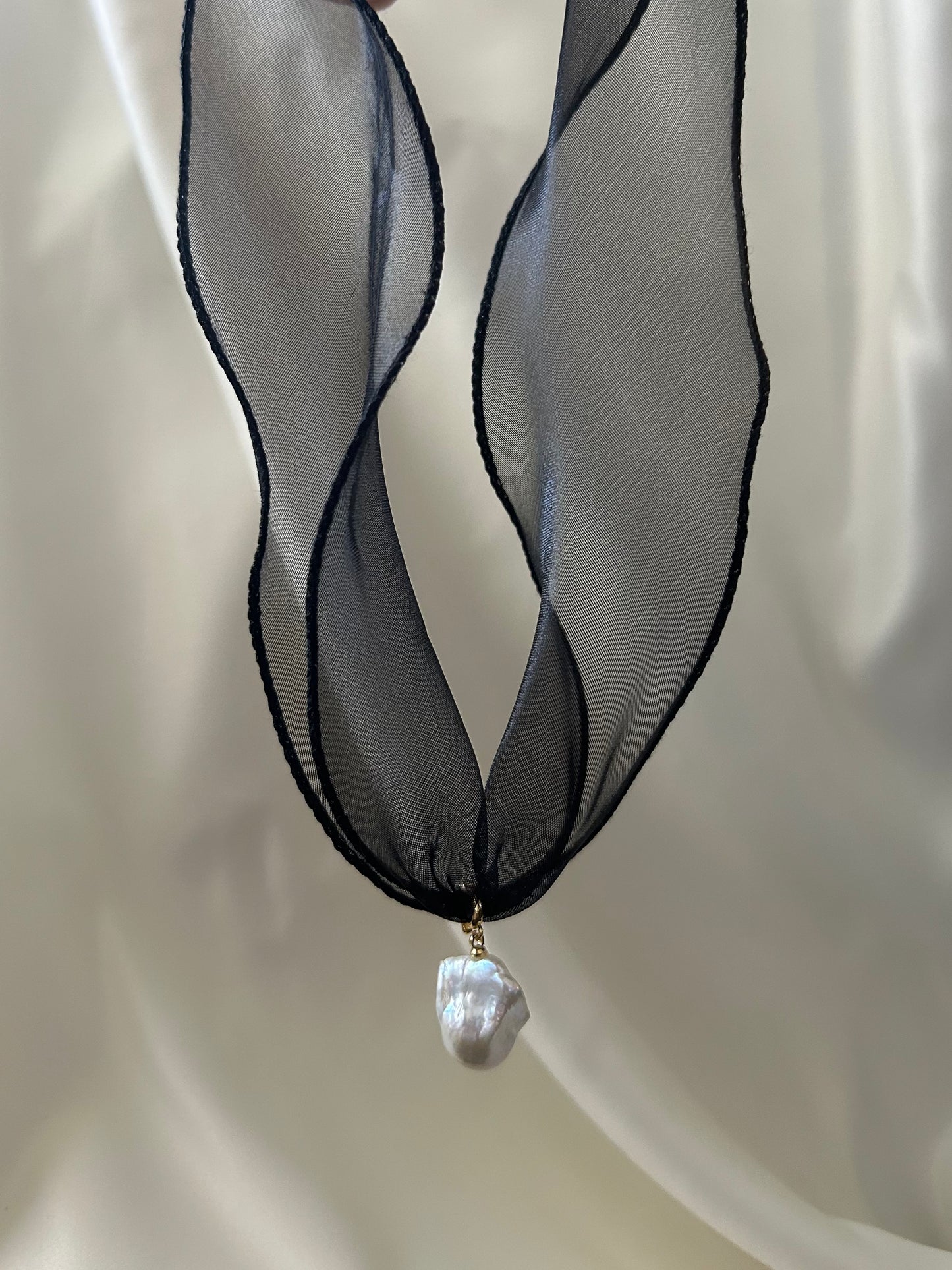 A black sheer ribbon necklace with a single irregular white baroque pearl pendant, displayed against a soft white draped fabric background.