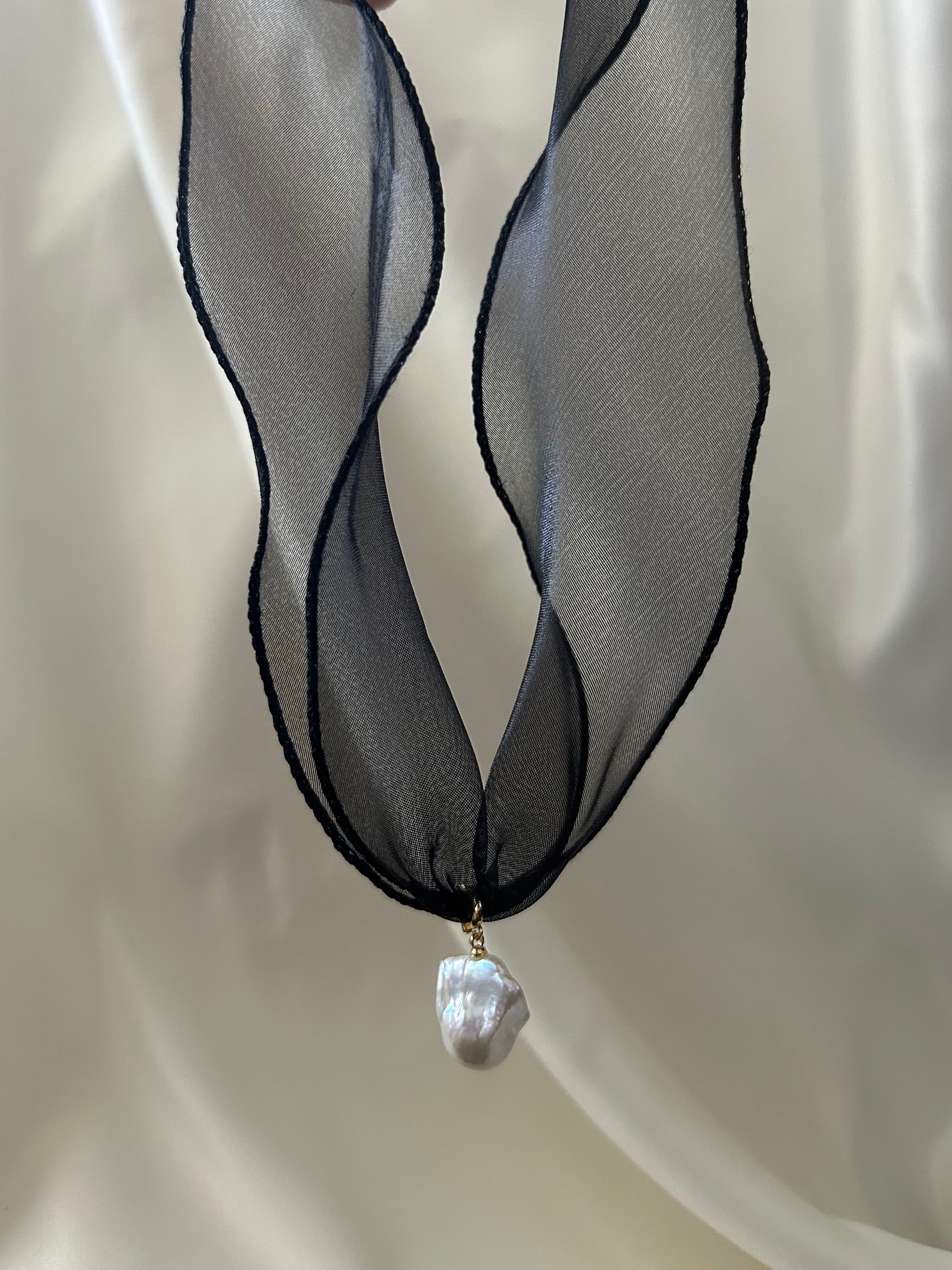 A black sheer ribbon necklace with a single irregular white baroque pearl pendant, displayed against a soft white draped fabric background.