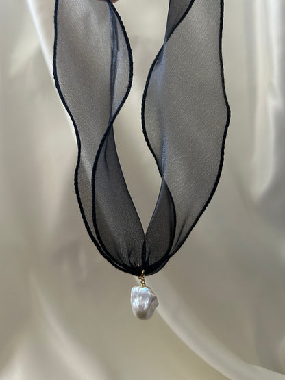 A black sheer ribbon necklace with a single irregular white baroque pearl pendant, displayed against a soft white draped fabric background.