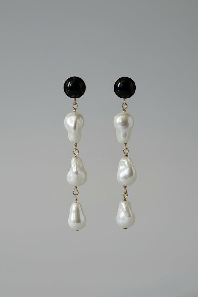 A pair of black drop earrings with irregular white pearl-like beads hanging from a black display stand against a draped cream satin fabric background.