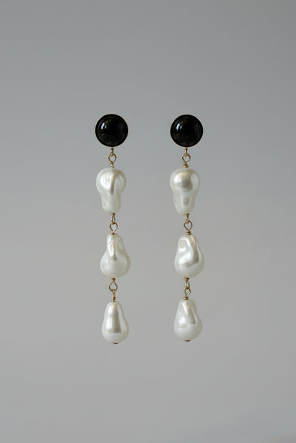 A pair of black drop earrings with irregular white pearl-like beads hanging from a black display stand against a draped cream satin fabric background.