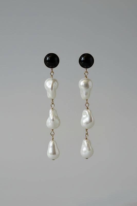 A pair of black drop earrings with irregular white pearl-like beads hanging from a black display stand against a draped cream satin fabric background.