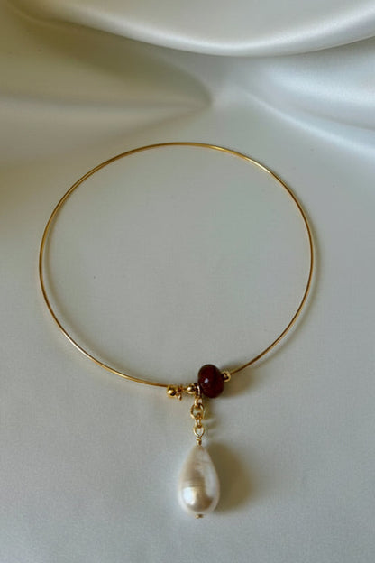 A delicate gold wire bangle bracelet featuring a small dark red bead, a gold spacer, and a dangling white teardrop pearl, displayed on draped white satin fabric.