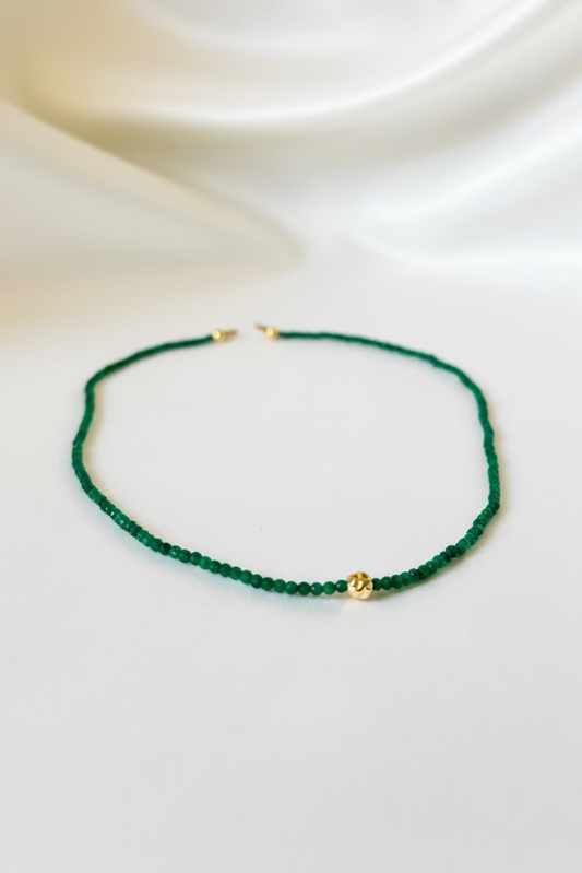 A delicate beaded necklace featuring dark green faceted beads and a small gold accent bead, displayed on a draped white satin fabric background.