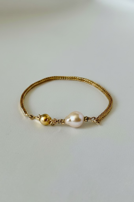 A delicate gold chain bracelet featuring a single baroque pearl and a gold bead accent, displayed on a clean, light gray surface.