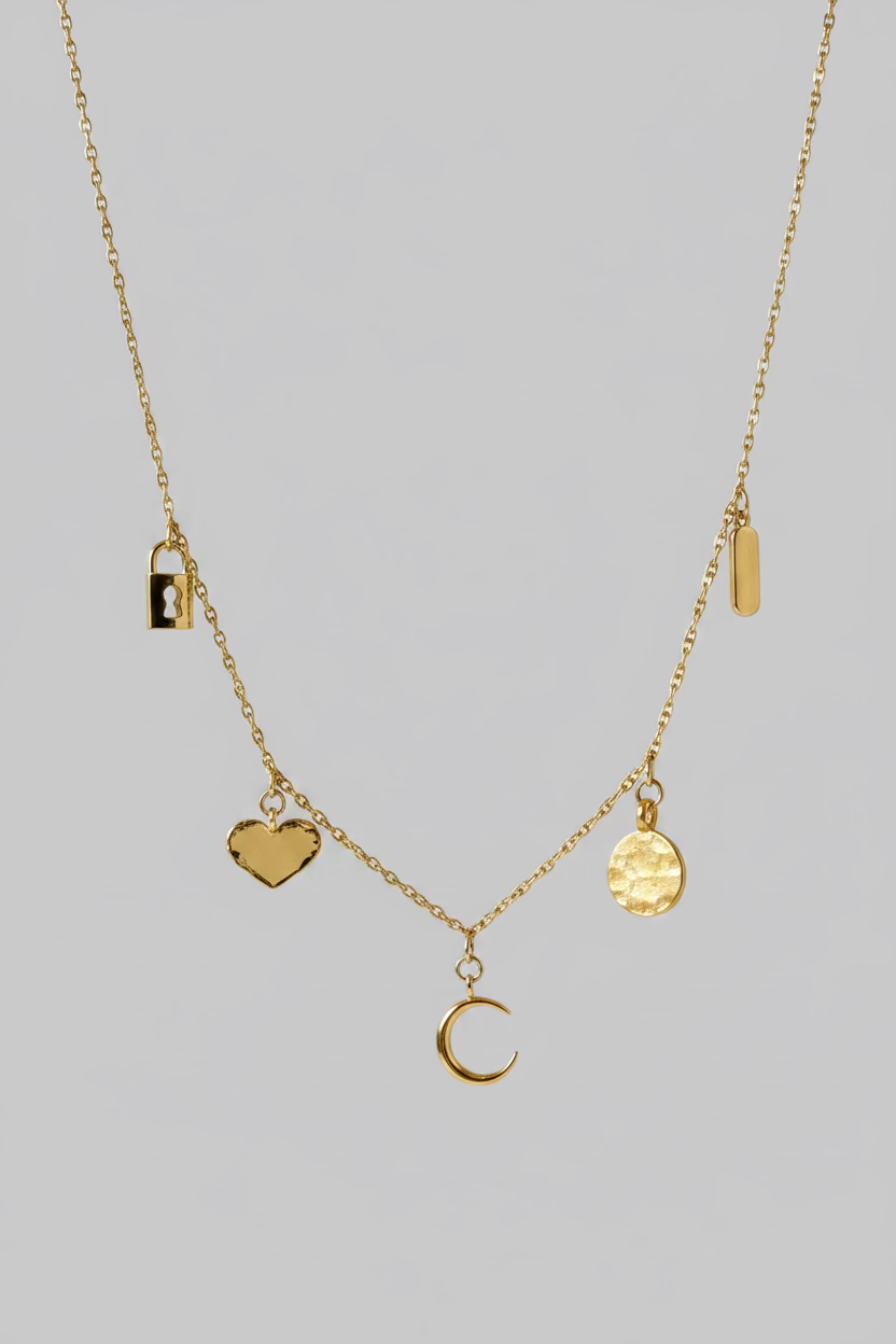 A delicate gold necklace displayed on a draped white satin fabric. The necklace features multiple small charms, including a crescent moon, a textured gold disc, a small rectangular charm, and a dark brown heart-shaped charm.