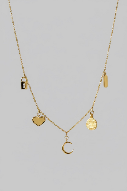 A delicate gold necklace displayed on a draped white satin fabric. The necklace features multiple small charms, including a crescent moon, a textured gold disc, a small rectangular charm, and a dark brown heart-shaped charm.