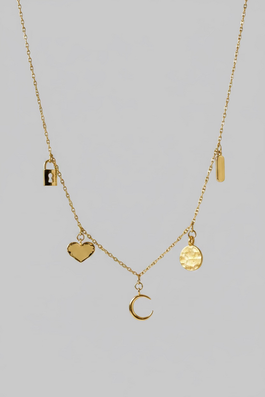 A delicate gold necklace displayed on a draped white satin fabric. The necklace features multiple small charms, including a crescent moon, a textured gold disc, a small rectangular charm, and a dark brown heart-shaped charm.
