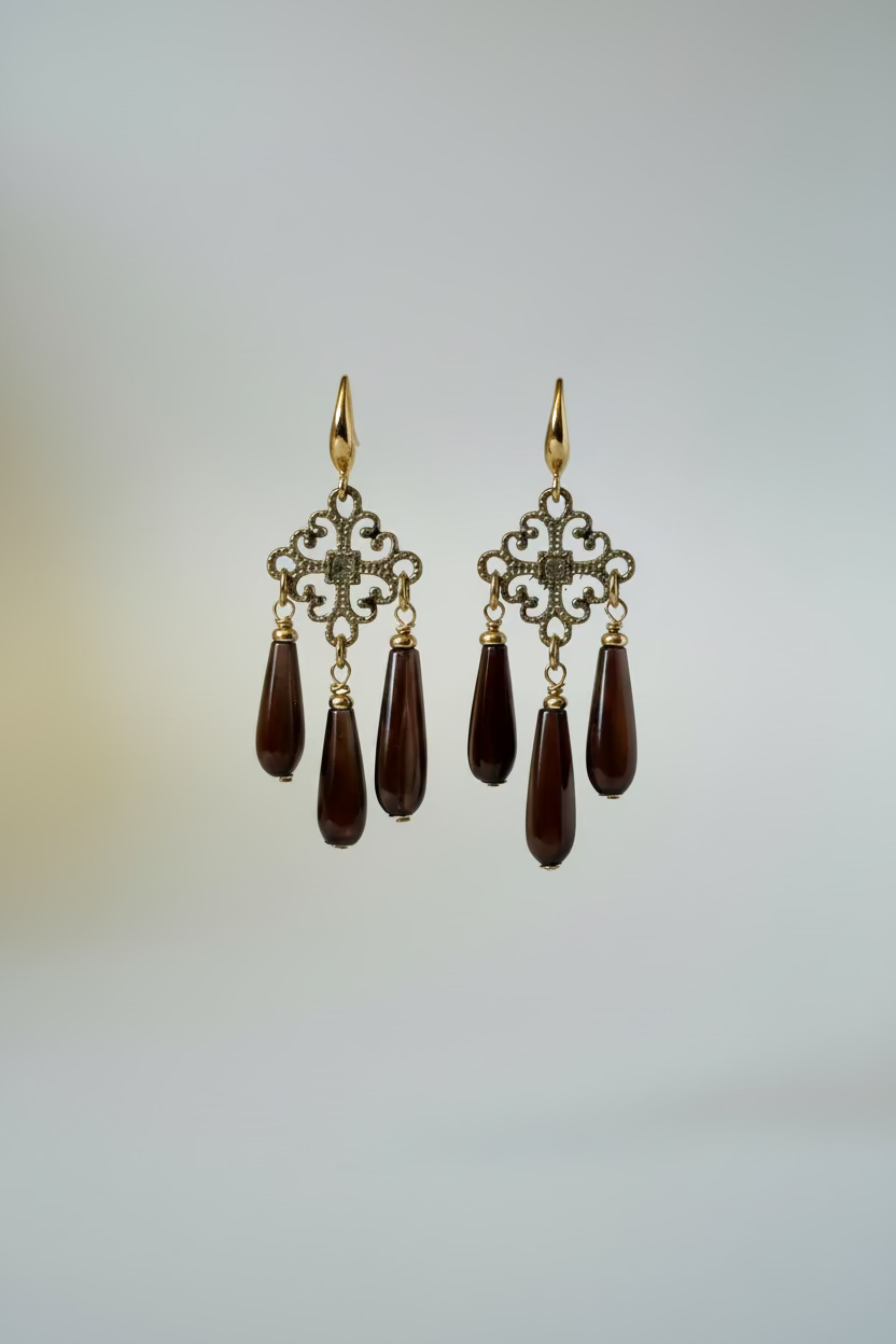 A pair of gold-tone dangle earrings with ornate filigree tops and three dark teardrop gemstone drops each, displayed on a metal stand against a draped white fabric background.