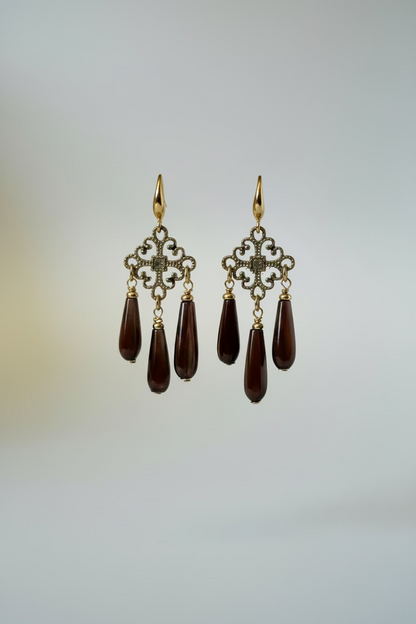 A pair of gold-tone dangle earrings with ornate filigree tops and three dark teardrop gemstone drops each, displayed on a metal stand against a draped white fabric background.