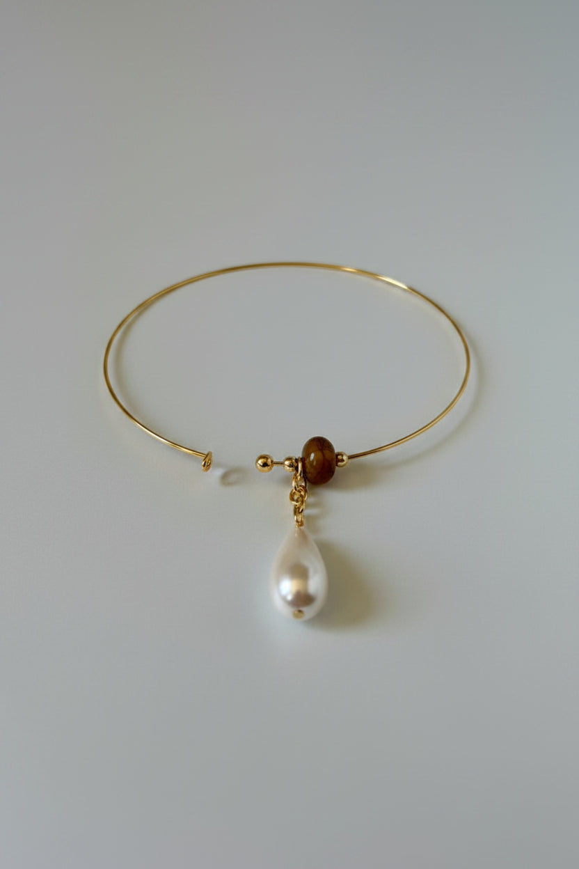 A delicate gold wire bangle bracelet featuring a small brown bead and a dangling white baroque pearl, displayed on a draped white satin fabric.