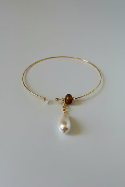 A delicate gold wire bangle bracelet featuring a small brown bead and a dangling white baroque pearl, displayed on a draped white satin fabric.
