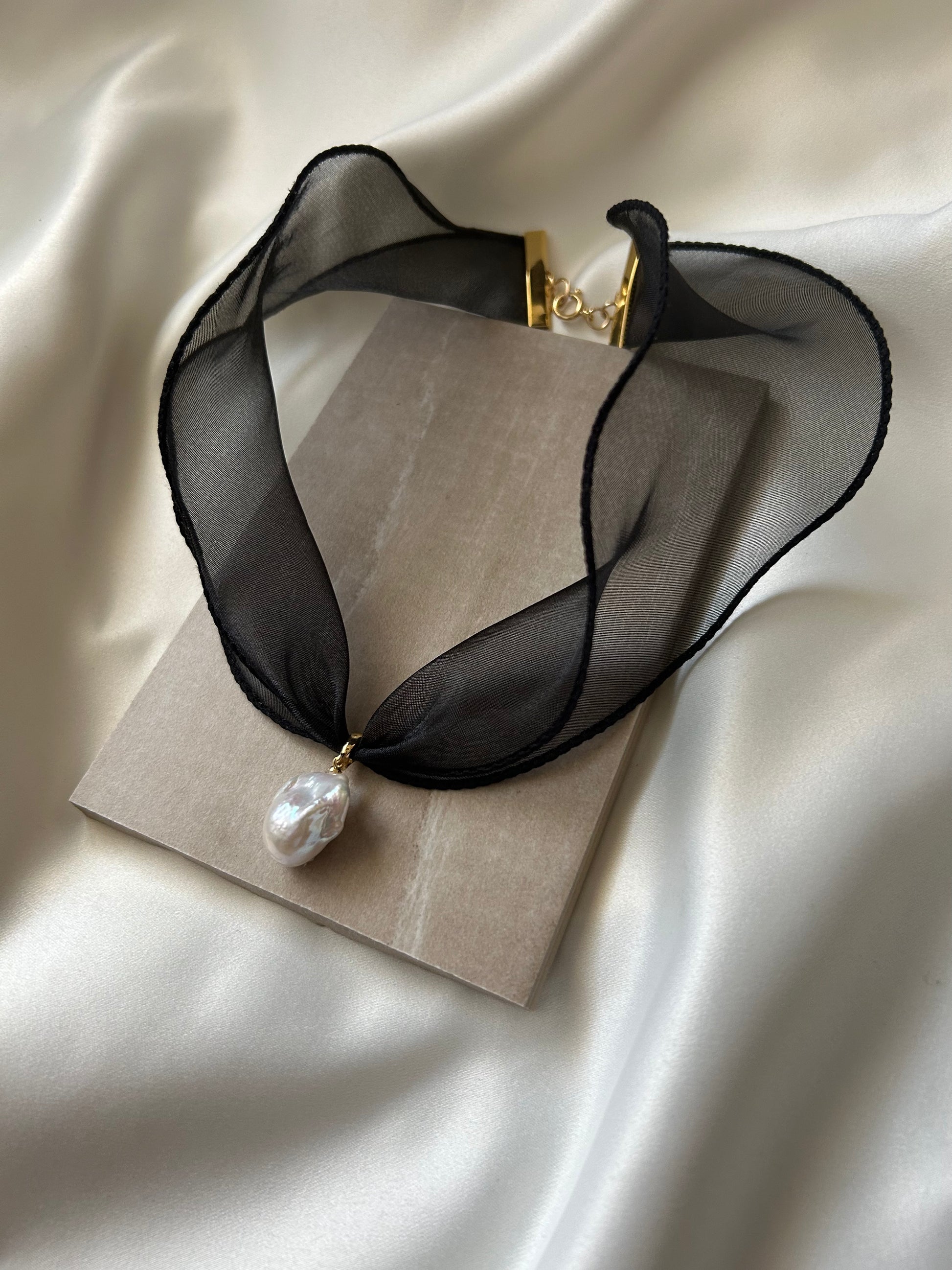 A black sheer mesh choker necklace with a gold clasp and a single white baroque pearl pendant, displayed on a beige card over a cream satin fabric background.