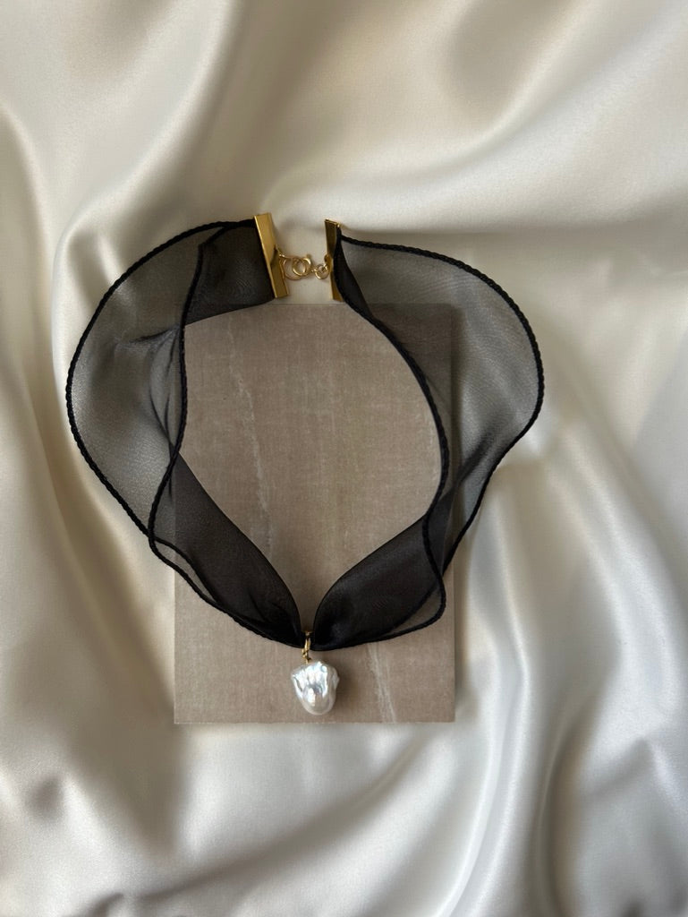 A black sheer organza collar necklace with a gold clasp and a single irregular white pearl pendant, displayed on a beige fabric stand against a draped cream satin background.