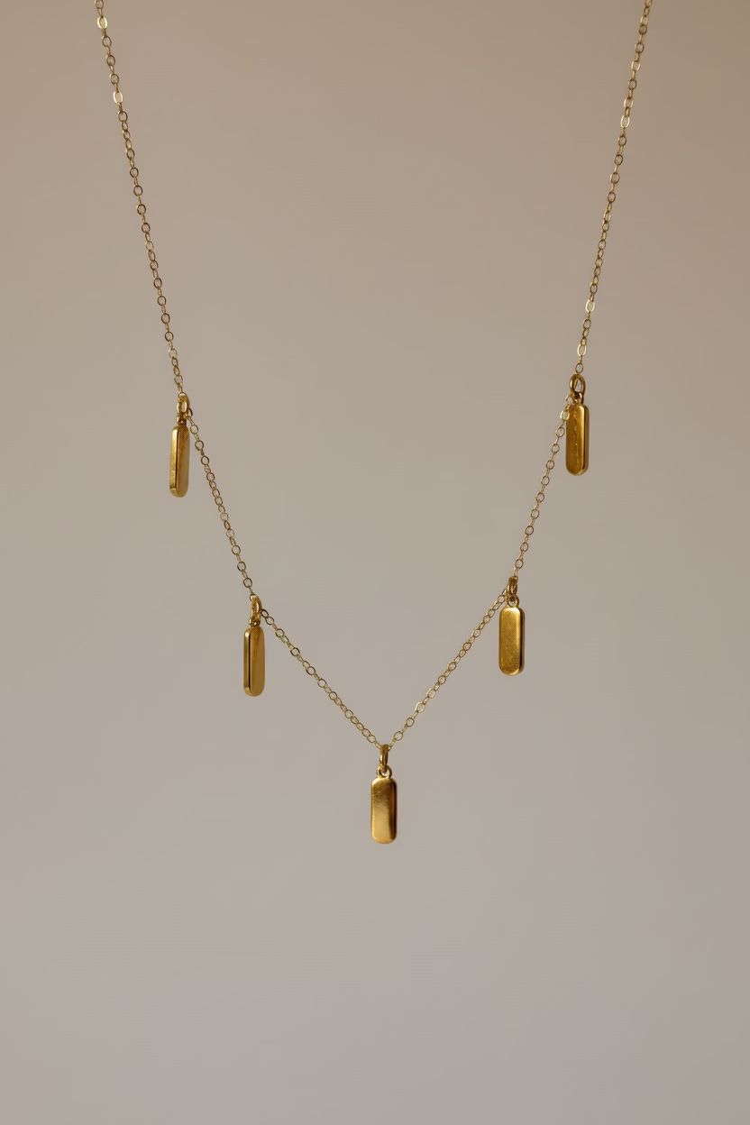 A delicate gold chain necklace with five small rectangular gold pendants, draped elegantly over a draped white satin fabric background.