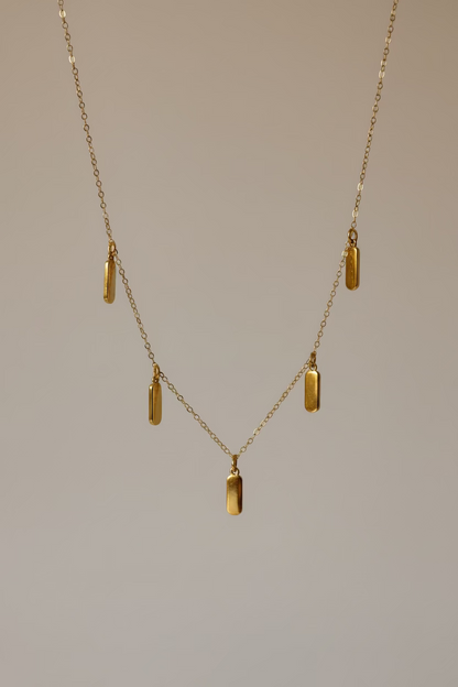 A delicate gold chain necklace with five small rectangular gold pendants, draped elegantly over a draped white satin fabric background.
