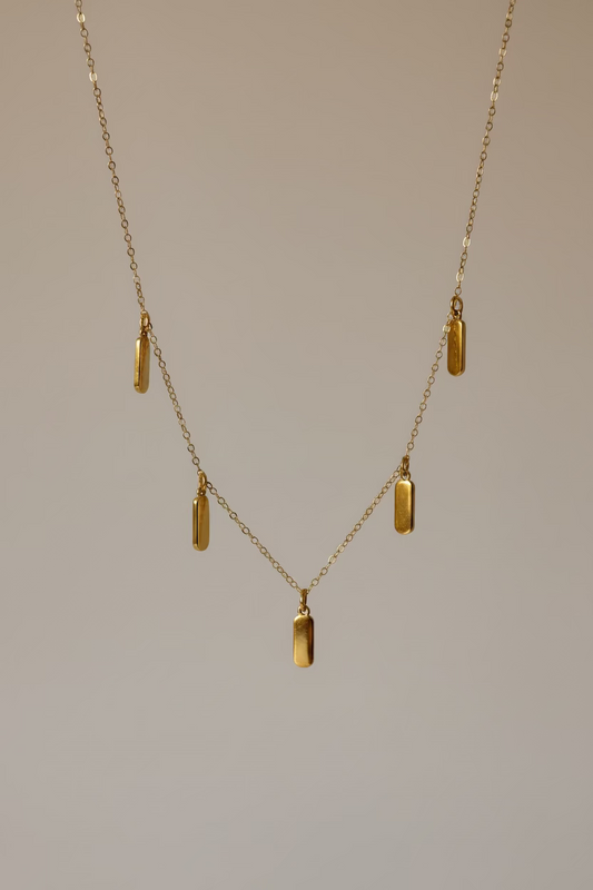 A delicate gold chain necklace with five small rectangular gold pendants, draped elegantly over a draped white satin fabric background.
