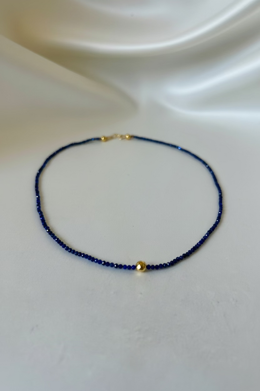 A delicate beaded necklace with deep blue beads and small gold accents, displayed on a draped white satin fabric background.