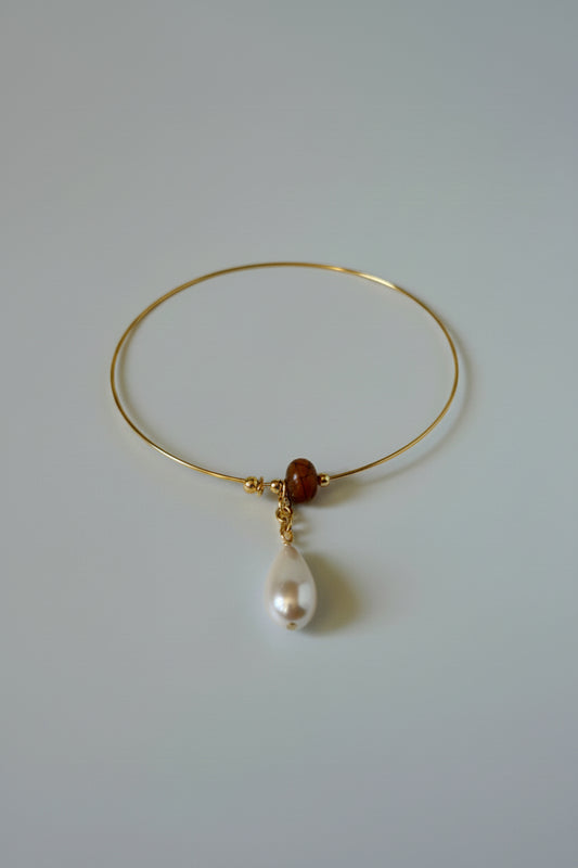 A delicate gold wire bangle bracelet featuring a small dark brown bead, a gold chain, and a dangling white freshwater pearl, displayed on a draped white satin fabric.