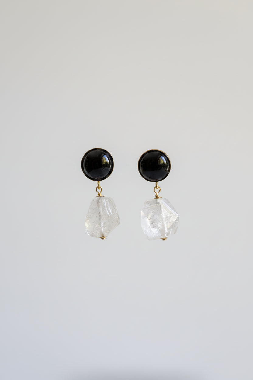A pair of black stud earrings with faceted clear crystal dangles, displayed on a black stand against a softly lit beige and yellow background.