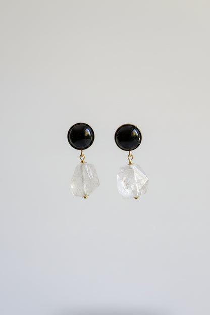 A pair of black stud earrings with faceted clear crystal dangles, displayed on a black stand against a softly lit beige and yellow background.
