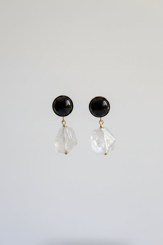 A pair of black stud earrings with faceted clear crystal dangles, displayed on a black stand against a softly lit beige and yellow background.