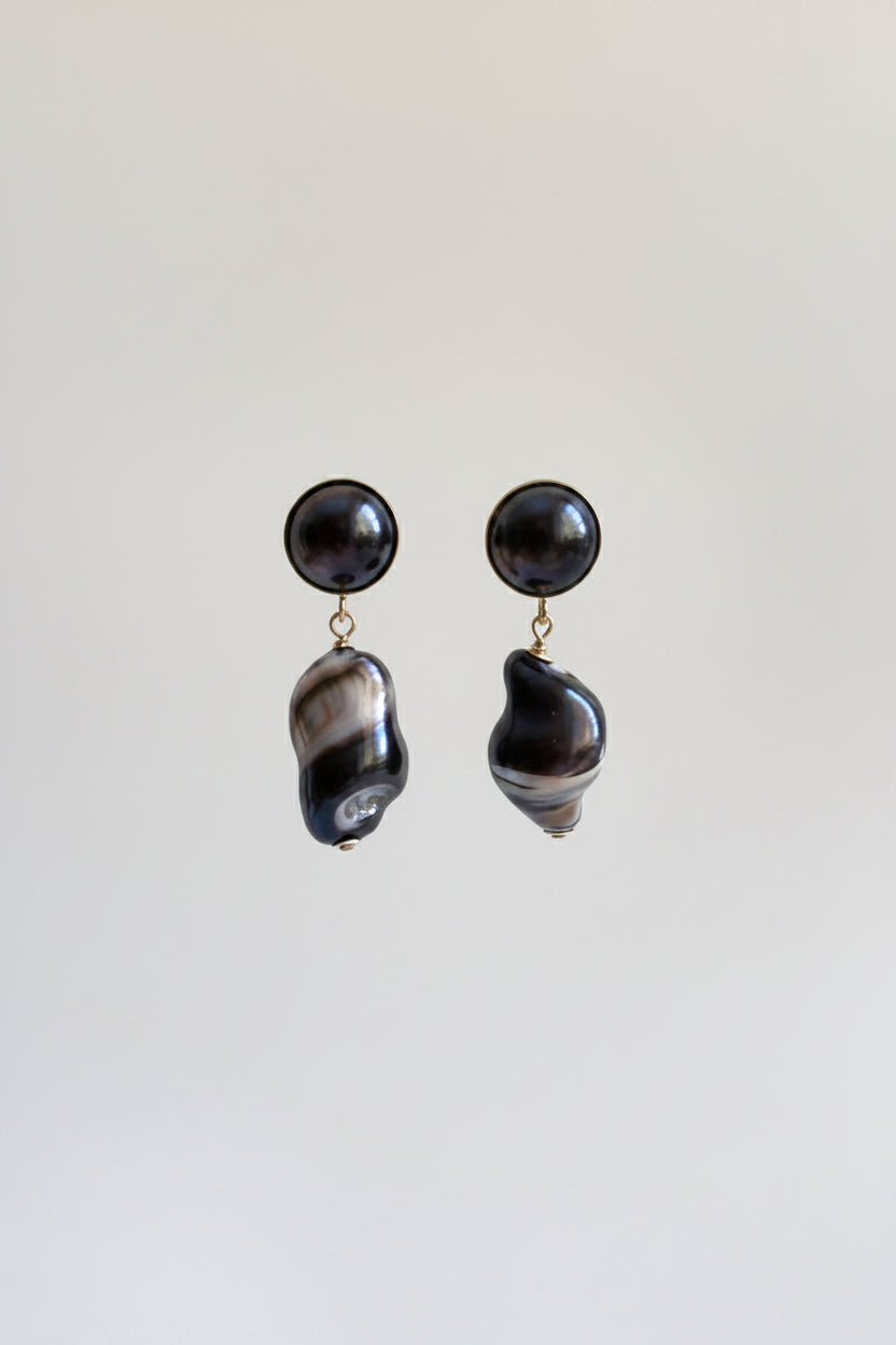 A pair of statement dangle earrings featuring round black pearl studs and irregular baroque shell drops with a black and white marbled pattern, suspended against a plain light gray background.