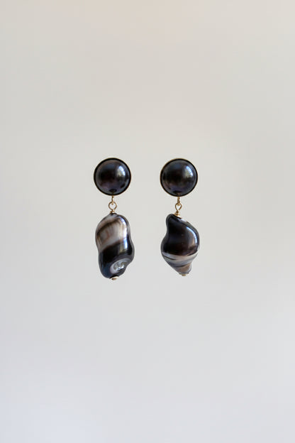 A pair of statement dangle earrings featuring round black pearl studs and irregular baroque shell drops with a black and white marbled pattern, suspended against a plain light gray background.