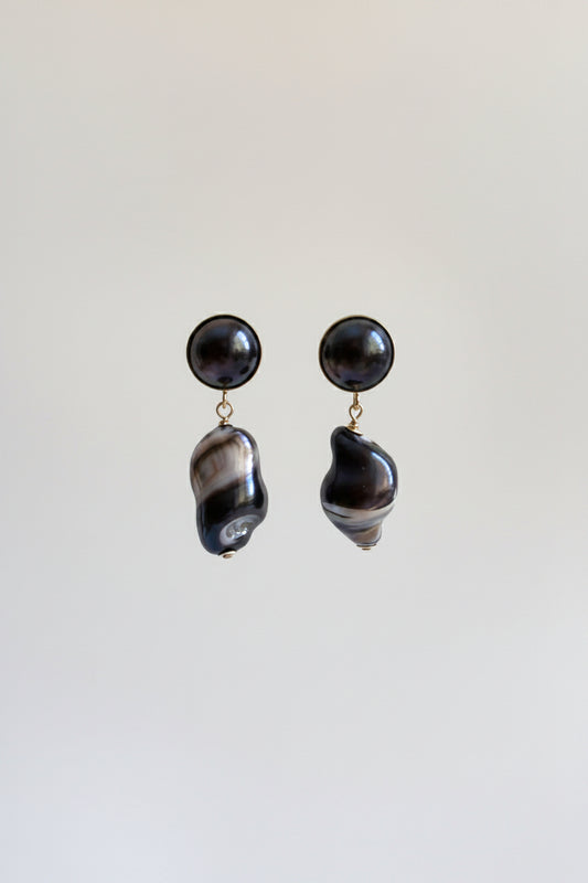 A pair of statement dangle earrings featuring round black pearl studs and irregular baroque shell drops with a black and white marbled pattern, suspended against a plain light gray background.