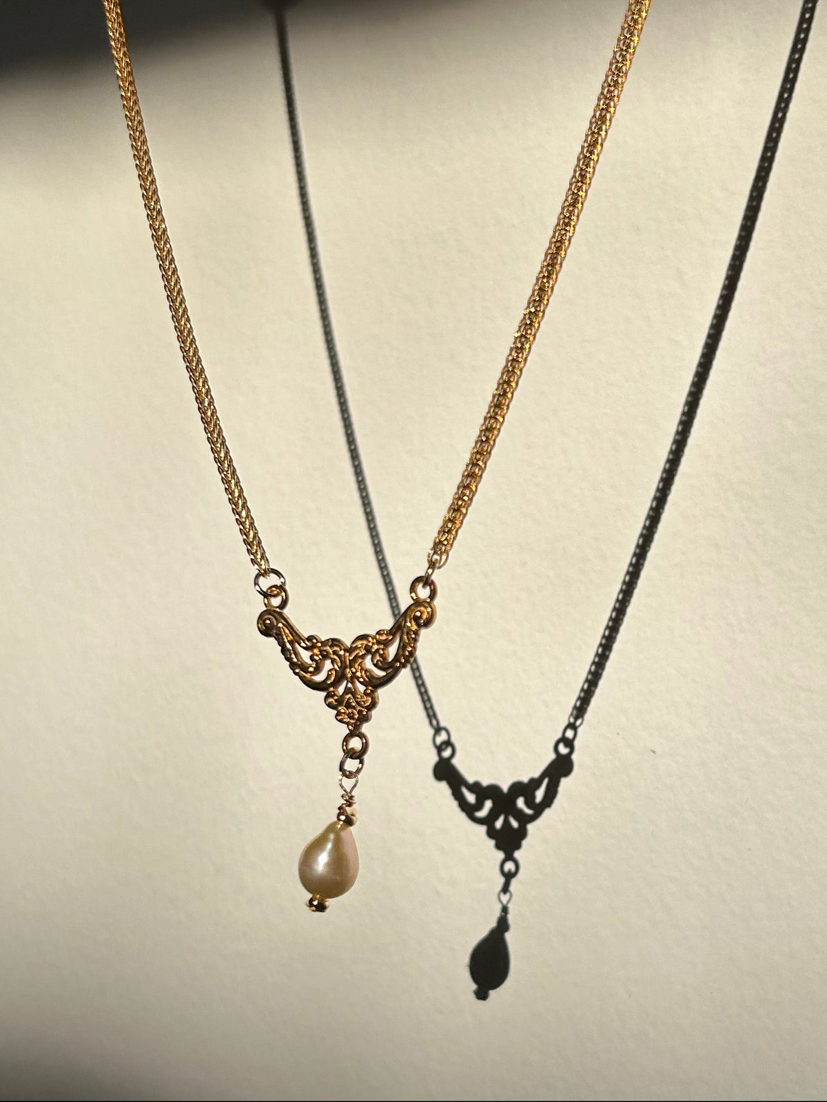 Two delicate necklaces displayed against a plain light background: one gold-toned chain necklace with an ornate filigree pendant and a small pearl drop, and one black-toned chain necklace with a matching filigree pendant and teardrop shadow.