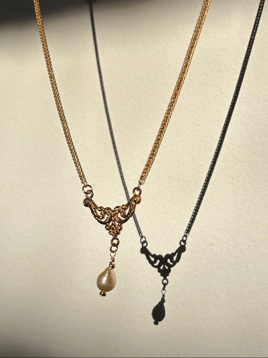 Two delicate necklaces displayed against a plain light background: one gold-toned chain necklace with an ornate filigree pendant and a small pearl drop, and one black-toned chain necklace with a matching filigree pendant and teardrop shadow.